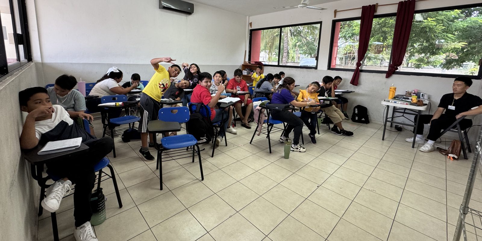 Students at desks during a morning math session in a Yucatán classroom, with a BTST volunteer seated at the side